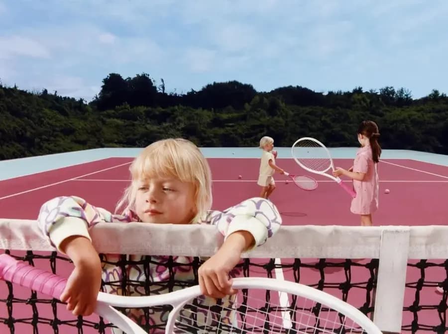 A children's editorial production on a pink tennis court surrounded by green hills.